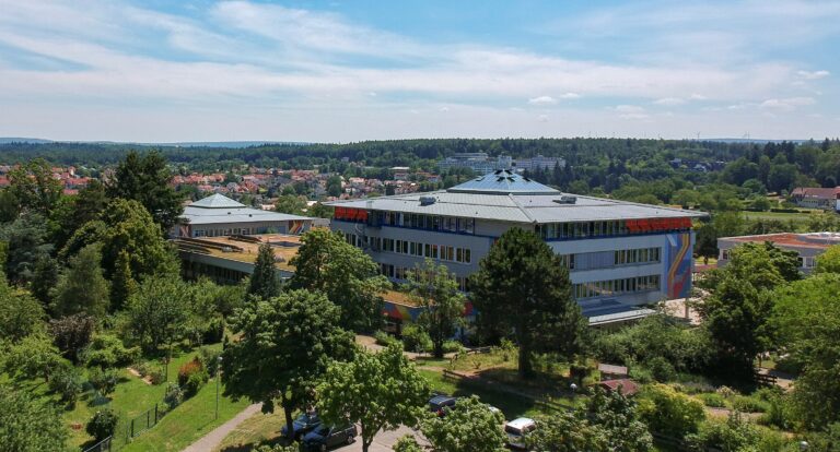 Das Bild zeigt eine Außenansicht eines modernen Schulgebäudes, das von Bäumen und Grünflächen umgeben ist. Das Gebäude hat mehrere Etagen und eine auffällige, flache Dachkonstruktion mit einem zentralen, kuppelförmigen Element. Die Fassade ist hell und hat große Fenster, die viel Licht ins Innere lassen. An einer Seite des Gebäudes sind bunte, geometrische Muster zu sehen, die dem Gebäude eine lebendige Note verleihen. Im Vordergrund sind verschiedene Bäume und Sträucher zu erkennen, die eine grüne Umgebung schaffen. Im Hintergrund sind weitere Gebäude und eine hügelige Landschaft sichtbar, die den Eindruck einer ländlichen Umgebung vermitteln. Der Himmel ist blau mit einigen Wolken, was auf einen sonnigen Tag hinweist.