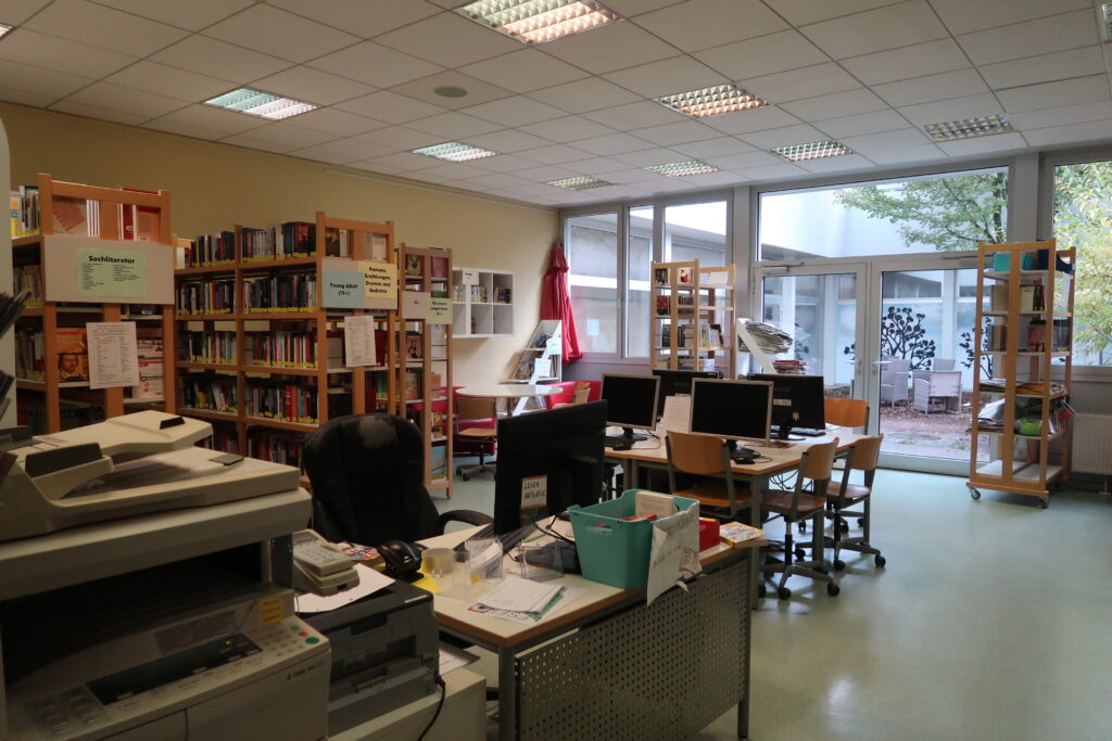 Das Bild zeigt einen Bibliotheksraum. Mehrere Bücherregale, die mit Büchern gefüllt sind, stehen entlang der Wände. Im Raum befinden sich außerdem Schreibtische mit Computern und verschiedenen Büroartikeln. Im Vordergrund links steht ein Drucker oder Kopierer. Am hinteren Ende des Raumes gibt es große Fenster und Glastüren, durch die Tageslicht hereinfällt. Draußen ist ein Außenbereich zu erkennen. Die Decke ist mit Leuchtstofflampen ausgestattet.