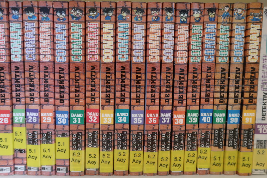 Das Bild zeigt eine Reihe von Manga-Büchern, die nebeneinander in einem Regal stehen. Es handelt sich um die Serie „Detektiv Conan“ von Gosho Aoyama. Auf jedem Buchrücken sind folgende Elemente zu sehen: Der Serientitel „Detektiv Conan“ Die jeweilige Bandnummer (z. B. 26 bis 40, dann 89 bis 91) Ein kleines Porträt des Hauptcharakters oben auf dem Buchrücken Unter der Bandnummer steht jeweils ein Kürzel wie „5.1 Aoy“ oder „5.2 Aoy“ Die Bücher sind ordentlich nebeneinander aufgereiht und bilden eine farblich einheitliche Reihe.