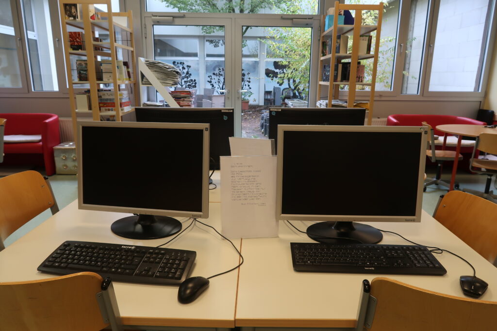 Auf einem weißen Tisch stehen zwei Computerbildschirme nebeneinander, jeweils mit Tastatur und Maus davor. Zwischen den Monitoren liegt ein Blatt Papier mit handgeschriebenem Text, der auf dem Bild jedoch nicht lesbar ist. Im Hintergrund befinden sich Bücherregale, die mit Büchern gefüllt sind. Davor stehen rote Stühle. Der Raum hat große Fenster, durch die Tageslicht einfällt. Draußen ist grüne Vegetation zu sehen.