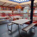 Das Bild zeigt einen Innenraum mit hoher Decke und einem großen Oberlicht, durch das Tageslicht einfällt. Der Boden ist blau, darauf stehen mehrere Picknicktische mit Bänken. Die Fläche ist von roten Geländern umgeben. Auf der rechten Seite führt eine Treppe nach unten zu einer tiefer gelegenen Ebene. Im Hintergrund hängt eine bunte, zeltartige Struktur von der Decke. Darunter befinden sich Dekorationen. An den Wänden im Hintergrund sind verschiedene Poster und Kunstwerke angebracht.