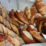 Das Bild zeigt eine Auswahl an Backwaren, die auf einem weißen Papiertuch in einem Behälter oder Korb liegen. Zu sehen sind verschiedene Brezeln – sowohl klassische als auch gedrehte Varianten. Einige sind mit grobem Salz bestreut. Daneben liegen längliche Brotstangen, teilweise mit Körnern oder Käse überbacken. Im Hintergrund befinden sich Croissants, von denen einige ebenfalls mit Käse bestreut sind.