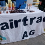 Das Bild zeigt einen Tisch im Freien, der auf einem gepflasterten Untergrund steht. Der Tisch ist mit einem weißen Tuch bedeckt. Auf dem Tuch steht in großen schwarzen Buchstaben „Fairtrade AG“. Auf dem Tisch sind verschiedene Produkte ausgestellt: mehrere Tafeln Schokolade, zwei Flaschen mit orangem Saft, einige verpackte Lebensmittel sowie eine blaue Tasche. Es liegen auch kleinere Gegenstände auf dem Tisch, deren Details nicht genau erkennbar sind.