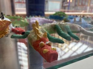 Das Bild zeigt eine Nahaufnahme eines bunten, handgefertigten Drachenmodells aus Ton. Der Drache hat einen roten Kopf mit gelben Augen und goldenen Stacheln auf dem Kopf. Sein Körper ist grün, ebenfalls mit goldenen Stacheln versehen, und er hat grüne Beine. Das Modell steht auf einer Glasoberfläche. Im Hintergrund sind weitere ähnliche Tonfiguren zu sehen, was darauf hindeutet, dass es sich um eine Ausstellung oder ein Schulprojekt handeln könnte. Das Bild hebt die kreative und handwerkliche Gestaltung der Figuren hervor.