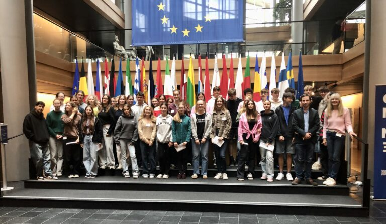 Das Bild zeigt eine Gruppe von Schüler:innen, die in einem großen, hellen Raum stehen. Im Hintergrund sind zahlreiche Flaggen der europäischen Länder aufgereiht, die in verschiedenen Farben leuchten. Über den Flaggen hängt eine große europäische Flagge mit einem blauen Hintergrund und gelben Sternen. Die Schüler:innen stehen auf einer Treppe und lächeln in die Kamera. Sie tragen unterschiedliche Kleidung, die von lässig bis sportlich reicht. Die Gruppe besteht aus Jungen und Mädchen unterschiedlichen Alters. Der Raum hat große Fenster, die viel Tageslicht hereinlassen, und die Wände sind aus Holz und Glas, was eine moderne Atmosphäre schafft.
