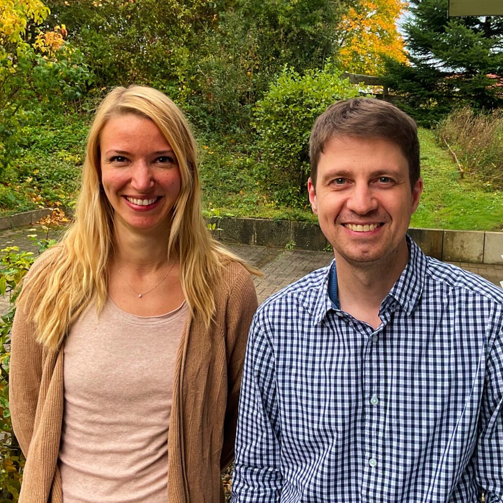 Das Bild zeigt zwei Personen, die nebeneinander stehen und in die Kamera lächeln. Die linke Person ist eine Frau mit langen, blonden Haaren, die leicht gewellt sind. Sie trägt ein helles Oberteil und eine beigefarbene Strickjacke. Ihr Gesichtsausdruck ist freundlich und offen. Die rechte Person ist ein Mann mit kurzen, braunen Haaren. Er trägt ein kariertes Hemd in Blau und Weiß. Auch er lächelt und wirkt freundlich. Im Hintergrund sind grüne Pflanzen und Bäume zu sehen, die auf eine natürliche Umgebung hinweisen. Der Boden ist gepflastert und es gibt einige Sträucher. Die Farben im Hintergrund sind überwiegend grün mit einigen herbstlichen Farbtönen.