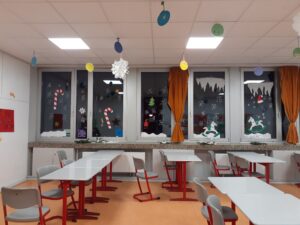 Das Bild zeigt einen hellen Raum, der als Klassenzimmer oder Aufenthaltsraum genutzt wird. An den Fenstern sind verschiedene weihnachtliche Dekorationen angebracht. Dazu gehören Schneeflocken, Zuckerstangen, Weihnachtsbäume und Figuren, die im Schnee spielen. Die Fenster sind mit weißen Vorhängen umrahmt, die orangefarben sind und zur Gemütlichkeit des Raumes beitragen. Im Raum stehen mehrere Tische mit grauen Tischplatten und roten Tischbeinen. Die Stühle sind ebenfalls grau mit roten Gestellen. Einige Stühle sind an den Tischen platziert, während andere leer sind. Die Decke ist hell und hat mehrere Deckenleuchten, die den Raum gut ausleuchten. Der Boden ist in einem warmen Orangeton gehalten, was eine freundliche Atmosphäre schafft.