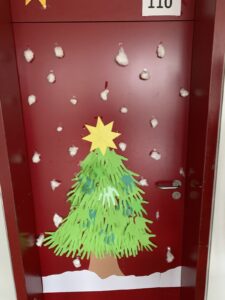 Das Bild zeigt die Tür eines Raumes, die in einem kräftigen Rot gestrichen ist. Oben an der Tür ist die Nummer "110" angebracht. In der Mitte der Tür ist ein großer, aus Papier gestalteter Weihnachtsbaum zu sehen, der aus grünen Handabdrücken besteht. Der Baum hat eine braune Stammfarbe und ist am Boden mit einer weißen Schneedecke aus Papier verziert. Oben auf dem Baum befindet sich ein gelbes, sternförmiges Element. Um den Baum herum sind kleine, weiße Wattebäusche angebracht, die wie Schneeflocken aussehen und an der Tür verteilt sind. Die gesamte Dekoration vermittelt eine festliche und weihnachtliche Atmosphäre.