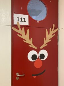 Das Bild zeigt die Tür eines Raumes, die in einem kräftigen Rot gestrichen ist. Oben an der Tür ist die Nummer "111" auf einem weißen Schild angebracht. In der Mitte der Tür sind große, runde Augen aus schwarzem und weißem Papier zu sehen, die einen freundlichen Gesichtsausdruck vermitteln. Darunter befindet sich eine rote Nase, die ebenfalls aus Papier gestaltet ist. Zusätzlich ist ein lächelnder Mund mit einem kleinen Zunge aus schwarzem Papier zu sehen. An den Seiten der Tür sind große, braune Geweih-Applikationen angebracht, die wie die Hörner eines Rentiers aussehen. Die gesamte Dekoration verleiht der Tür ein fröhliches und festliches Aussehen.