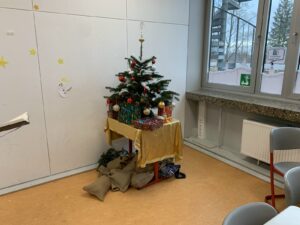 Das Bild zeigt eine Ecke eines Raumes, in der ein kleiner Weihnachtsbaum steht. Der Baum ist mit roten Kugeln, goldenen Ornamenten und einer silbernen Spitze geschmückt. Unter dem Baum befindet sich ein Tisch mit einer goldenen Tischdecke, auf dem mehrere Geschenke in buntem Geschenkpapier liegen. Neben dem Baum sind einige große, braune Säcke aus Jute, die auf dem Boden liegen. An der Wand hinter dem Baum sind einige gelbe Sterne und eine kleine, aus Papier ausgeschnittene Schneemannfigur angebracht. Das Fenster neben dem Baum zeigt eine verschneite Landschaft im Freien, und die Fensterbank ist mit einer grauen Steinplatte versehen. Der Boden des Raumes ist in einem warmen Orangeton gehalten, was eine einladende Atmosphäre schafft.