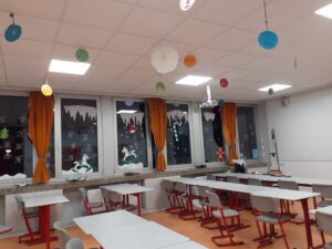 Das Bild zeigt einen festlich dekorierten Raum, der als Klassenzimmer genutzt wird. An den Fenstern hängen orangefarbene Vorhänge, die den Raum gemütlich wirken lassen. Die Fenster sind mit verschiedenen weihnachtlichen Motiven dekoriert, darunter Schneeflocken, eine Weihnachtsmütze und Figuren, die im Schnee spielen. Oben am Fenster sind große, weiße Schneeflocken aus Papier angebracht, die wie Eiszapfen aussehen. Von der Decke hängen bunte, runde Dekorationen in verschiedenen Farben, darunter Blau, Grün und Gelb. Im Raum stehen mehrere Tische mit grauen Tischplatten und roten Tischbeinen. Die Stühle sind ebenfalls grau mit roten Gestellen. Einige Tische sind leer, während andere Stühle an den Tischen platziert sind. In der Mitte des Raumes ist ein Beamer installiert, der an der Decke hängt. Der Boden ist in einem warmen Orangeton gehalten, was eine freundliche Atmosphäre schafft.
