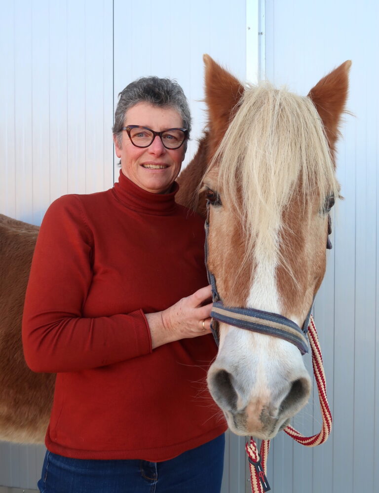 Das Bild zeigt eine Frau mit kurzen grauen Haaren und einer Brille, die ein rotes Turtleneck trägt. Sie steht neben einem Pferd, das eine braune und weiße Fellfarbe hat und mit einem Halfter ausgestattet ist. Die Frau lächelt und hält das Pferd am Halfter. Der Hintergrund ist ein hellblauer, glatter Untergrund, möglicherweise eine Wand oder ein Stall.
