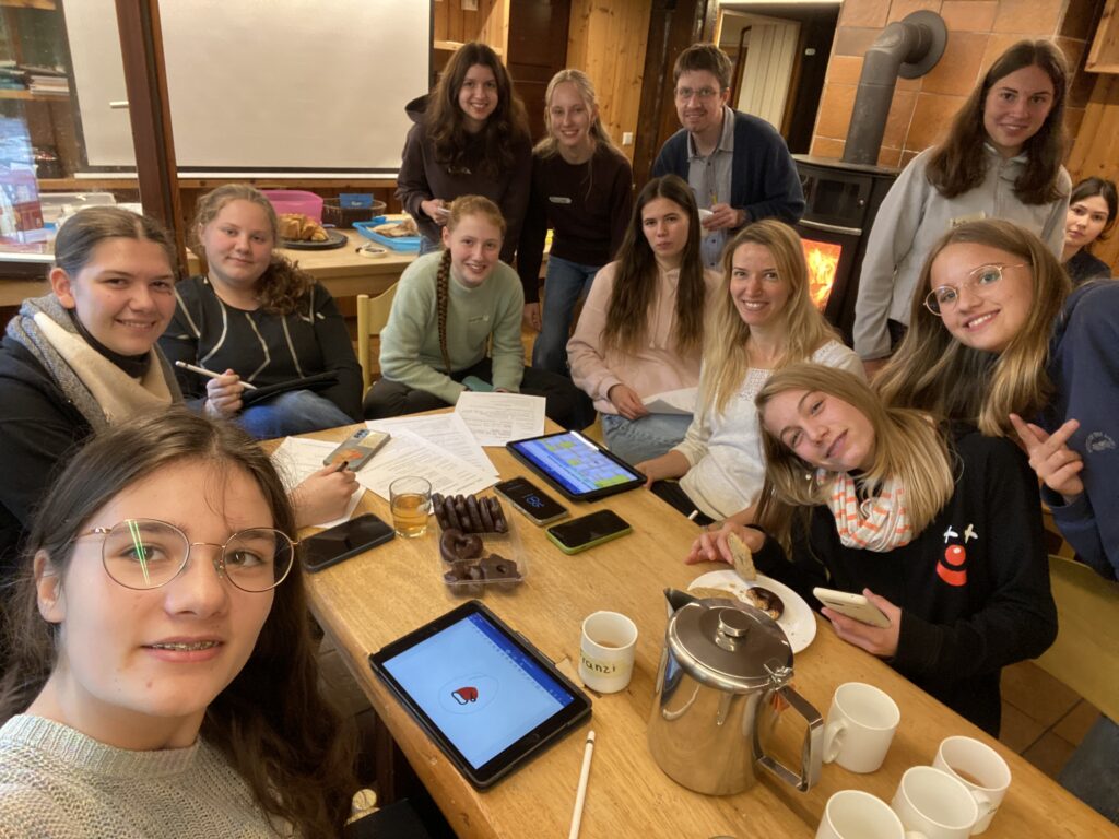 Mehrere Schülerinnen, eine Lehrerin und ein Lehrer sitzen und stehen um einen Tisch und lächeln in die Kamera. Auf dem Tisch liegen Handys, Arbeitsblätter, Tablets, gefüllte Becher, eine Teekanne und Lebkuchen. Im Hintergrund verschiedene Kuchen auf einem Tisch, und ein Kaminofen mit warmen Flammen.