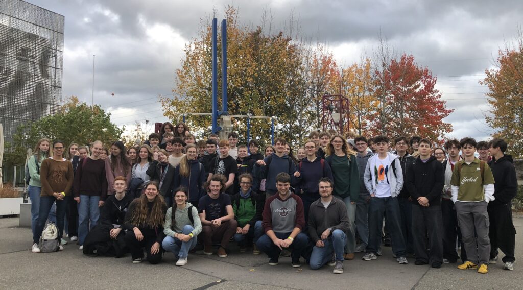 Eine Gruppe Schülerinnen und Schüler, im Hintergrund eine Skulptur und Bäume mit Herbstlaub, seitliche Gebäudeteile des Technorama.