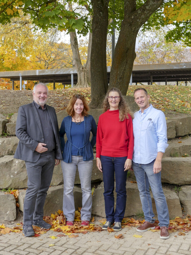 Vier Personen, zwei Männer und zwei Frauen, stehen vor einer Natursteinmauer. Im Im Hintergrund ein Baum sowie Dächer der Fahrradständer. Zu Füßen Herbstlaub.