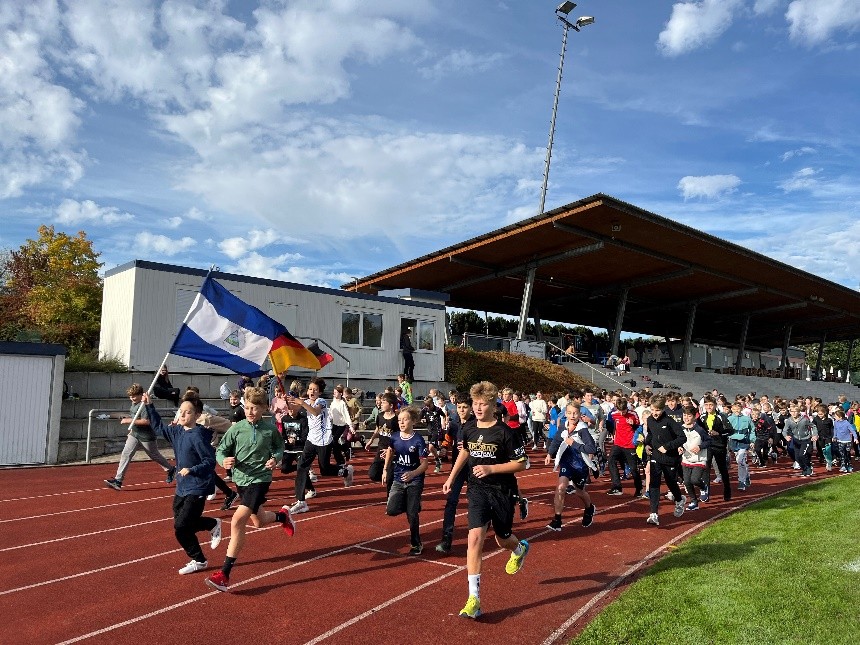 Ein Gruppe Schülerinnen und Schüler läuft im Stadion, im Hintergrund die Tribüne, vorne ein Junge, der eine Nicaragua Flagge schwenkt.