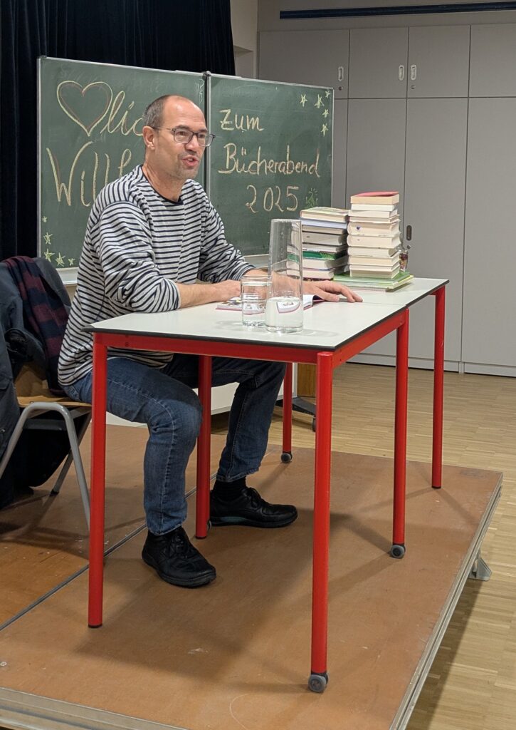 Ein Mann sitzend an einem Tisch auf dem diverse Bücher liegen, im Hintergrund eine beschriftete Tafel. Der Mann richtet Blick und Worte an ein Publikum, das nicht zu sehen ist.
