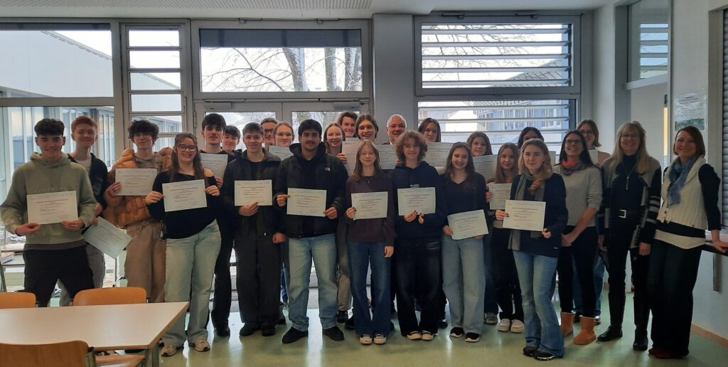rund 20 Schülerinnen und Schüler in einer Gruppe stehend, DELF Zertifikate in Händen. Dabei der Schulleiter und drei Französich Lehrerinnen. Im Hintergrund die Fensterwand der Mensa.