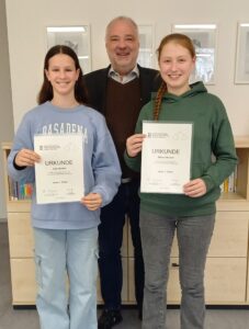 Zwei Schülerinnen stehend, in den Händen jeweils eine Urkunde haltend. Im Hintergrund mittgig stehend der Rektor. Alle lächelnd.