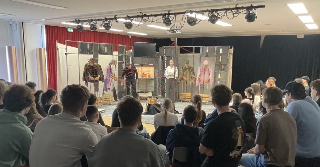 improvisierte Bühne im Klassenraum, 2 Schauspieler neben 4 "Puupen", vielen Aktenordnern, ein roter Faden, der kreuz und quer über die Bühne gespannt ist. im Vordergrund die Zuschauer, Schüler, die uns den Rücken zuwenden.