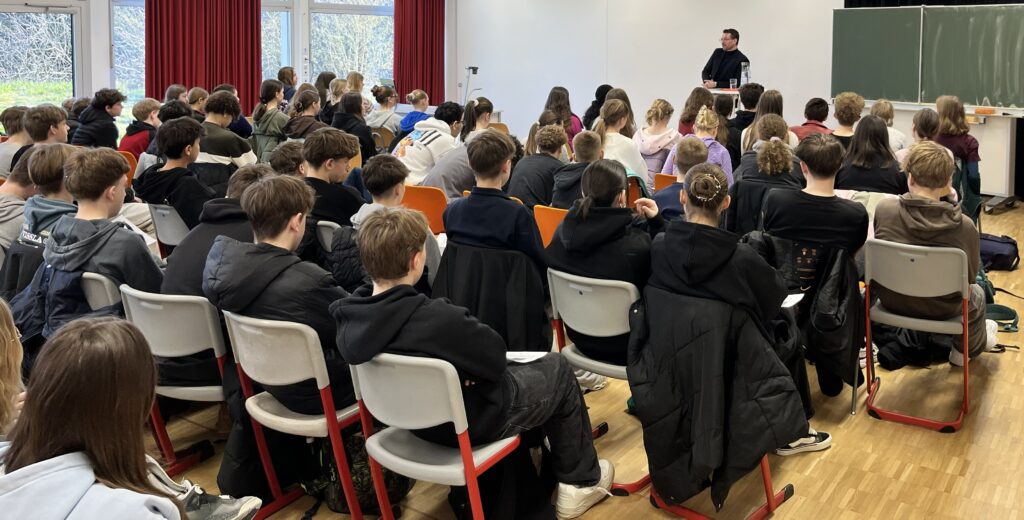 Bürgermeister, stehend am Pult vor großer Anzahl Schüler, diese sitzend, Hinterköpfe sichtbar.