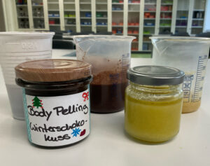 Auf einem Labortisch stehen zwei beschriftete Gläser mit selbstgemachtem Körperpeeling – eines dunkelbraun („Body Peeling Winterschokokuss“), eines gelblich. Dahinter befinden sich mehrere Messbecher und Becher mit Zutaten und Resten der Mischungen. Im Hintergrund sind Regale mit Materialkisten zu sehen.