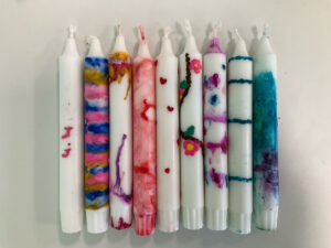 Neun Stabkerzen liegen nebeneinander auf hellem Untergrund. Einige sind weiß, andere bunt gestaltet: marmorierte und verlaufene Farben in Pink, Blau, Gelb, Türkis und Lila, dazu kleine Verzierungen wie Herzen, Punkte, Linien und Blumen. Jede Kerze hat ein individuelles Muster.