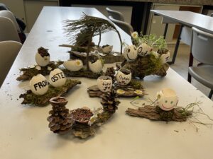 Auf einem Tisch stehen mehrere österliche Dekoarrangements aus bemalten Eierschalen. Die Schalen sind auf Rindenstücken mit Moos, Kiefernzapfen und kleinen Stöcken arrangiert, teils mit Gänseblümchen geschmückt oder mit Kressesprossen bepflanzt. Auf einigen Eiern steht „Frohe Ostern“, andere zeigen lachende Gesichter.