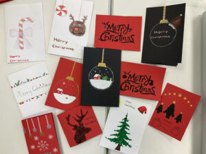 Mehrere selbst gestaltete Weihnachtskarten liegen ausgebreitet auf hellem Tisch. Die Karten in Rot, Weiß und Schwarz zeigen Motive wie Christbaumkugeln, Tannenbäume, Schneelandschaften, Zuckerstange und Rentier. Auf vielen steht „Merry Christmas“, teils in Handlettering oder mit Glitzer verziert. Jede Karte hat ein individuelles weihnachtliches Design.