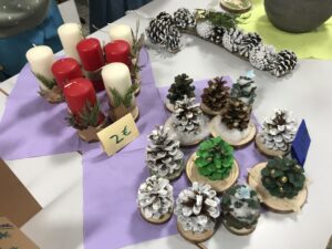 Auf einem Tisch stehen selbstgemachte Weihnachtsdekorationen. Links liegen rote und weiße Stumpenkerzen mit Tannengrün umwickelt, daneben ein Preisschild „2 €“. Rechts sind viele kleine Deko-Tannenbäume aus bemalten Tannenzapfen auf Holzscheiben arrangiert, teils mit Watte und goldenen Kugeln verziert. Im Hintergrund weitere Zapfenkränze.