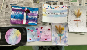 Mehrere selbst gestaltete Karten liegen auf Zeitungspapier. Zu sehen sind bunte Aquarelle in Blau, Lila, Rosa und Gelb, teils mit Schriftzügen wie „Für die weltbeste Mama“ oder „Hör nie auf zu träumen!“. Einige Karten haben aufgeklebte Deko wie kleine rote Herzen oder getrocknete Gräser und Blüten. Jede Karte ist individuell gestaltet.