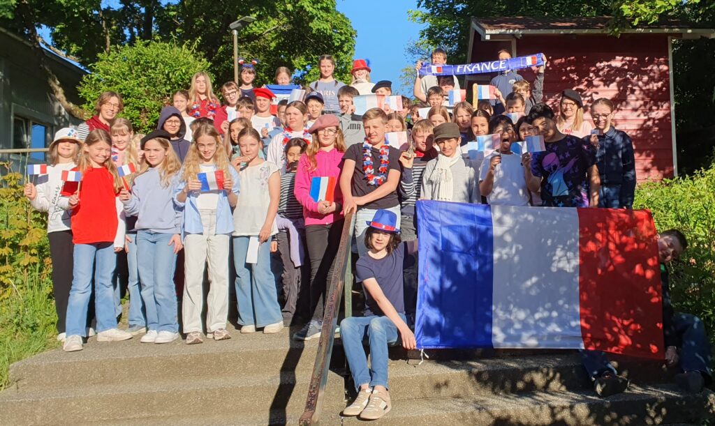 Das Bild zeigt eine große Gruppe von Kindern und Jugendlichen, die auf einer Außentreppe stehen und sitzen. Sie halten viele kleine französische Flaggen in den Händen und tragen teilweise Kleidung oder Accessoires in den Farben Blau, Weiß und Rot. Vor der Gruppe sitzt ein Kind und hält eine große französische Flagge, die teilweise Schatten von Bäumen zeigt. Im Hintergrund sind grüne Bäume und ein rotes Gartenhaus zu sehen. Die Stimmung wirkt fröhlich und gemeinschaftlich, das Wetter ist sonnig mit klarem blauem Himmel.