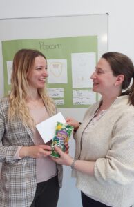 Zwei Lehrerinnen stehen sich gegenüber und lächeln sich an während ein Gewinnergeschenk mit Gutschein überreicht wird
