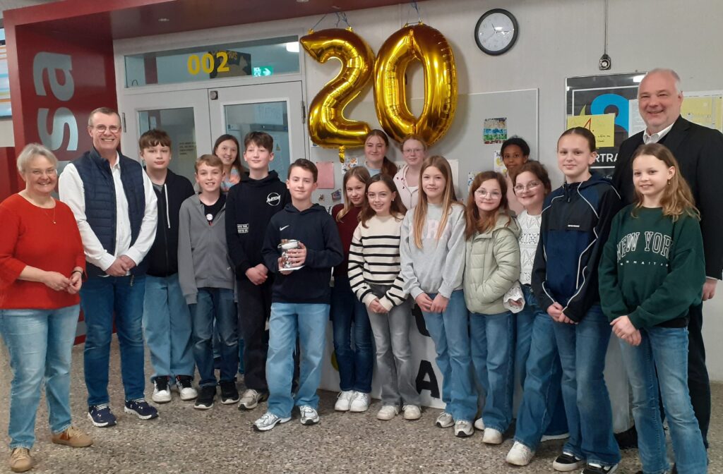 Schülergruppe mit 3 Lehrern, stehend. Mittig oberhalb der Gruppe eine goldene Jubiläums 20 (Ballone)