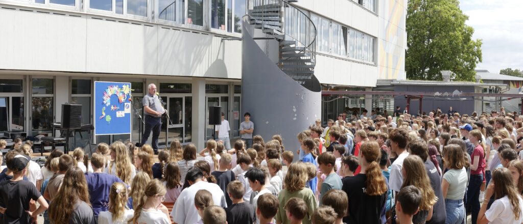 hunderte von Schülern verabschieden den Schulleiter, der auf einer Bühne steht, auf dem Schulhof vor dem Schulhaus
